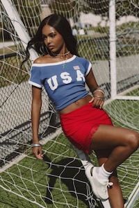 A person leans against a soccer goal net wearing a blue off‑shoulder top with “USA” lettering and a red skirt, styled with sneakers and jewelry.