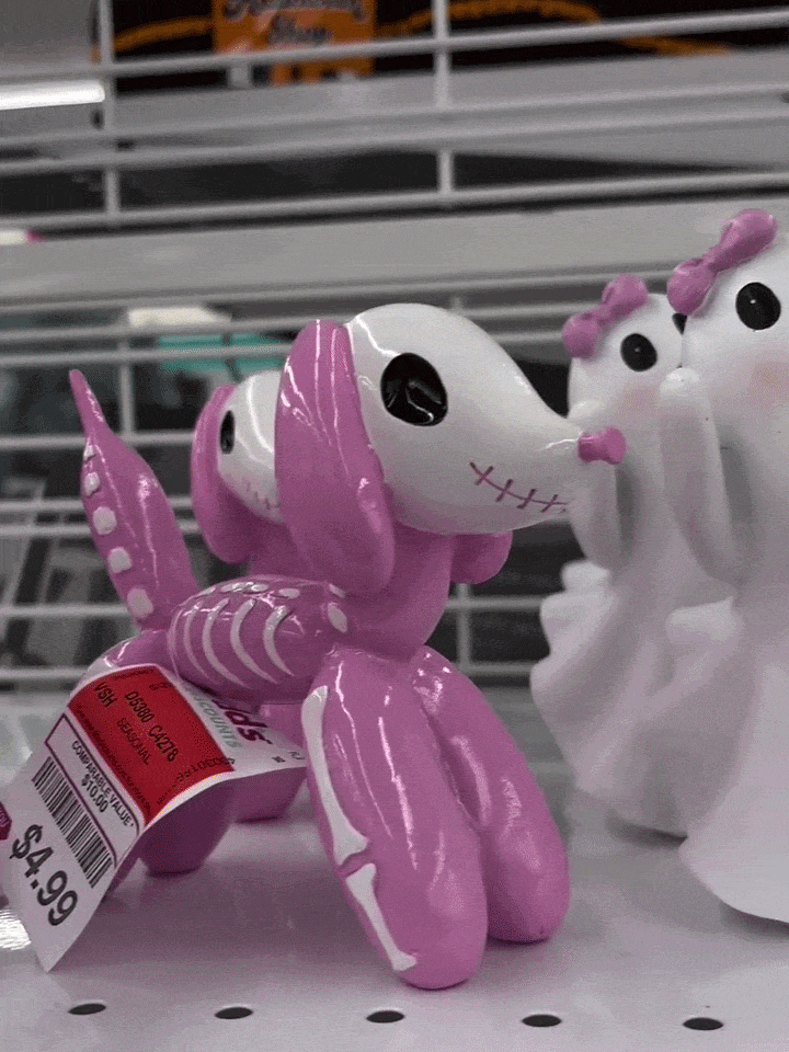 A pink dog figurine with skeleton details faces two white ghost figurines with black eyes, displayed taking a selfie on a retail shelf.