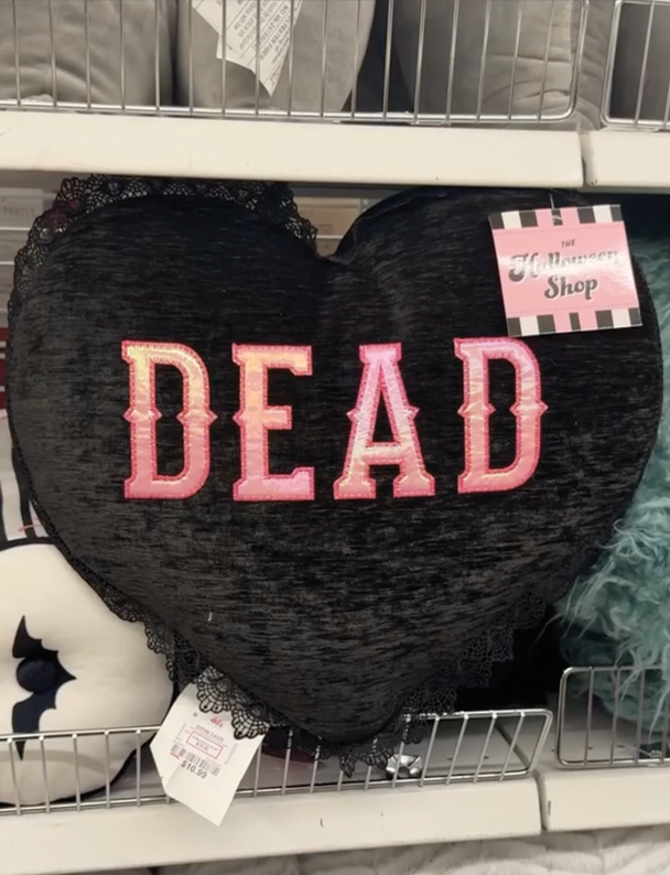 A black velvet-look heart-shaped decorative pillow featuring the word “DEAD” in large pink embroidered lettering, displayed on a store shelf.