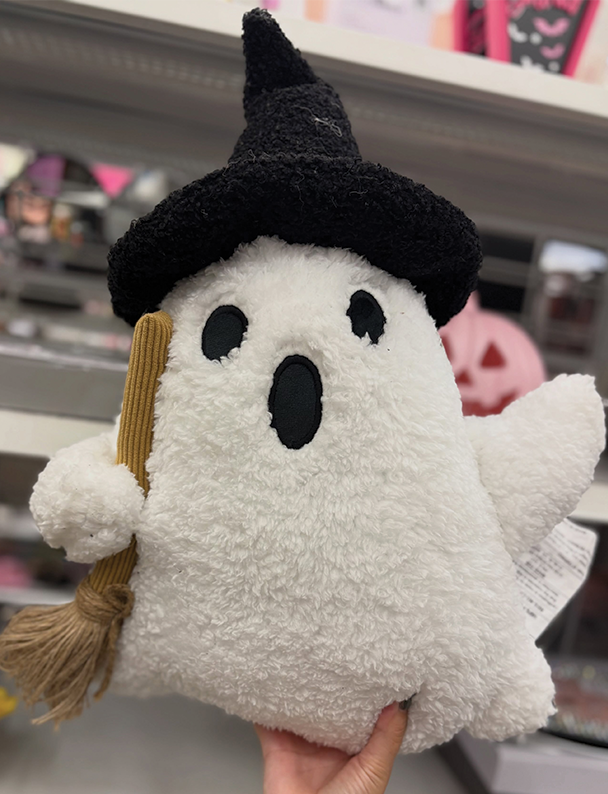 A fluffy white ghost pillow with black eyes and mouth, dressed in a black witch hat and holding a small broom, displayed in a store.