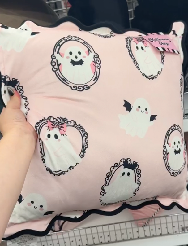 A square pink throw pillow decorated with playful ghost illustrations in ornate frames and finished with a black scalloped edge.