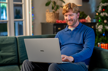 A parent siting on a couch in their living room doing their holiday shopping on a Dell laptop computer.