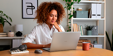 Woman in a home office working on a Dell laptop with a wireless mouse and headphones next to her.