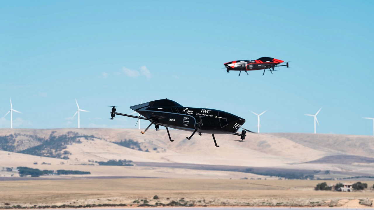 Two flying drones above a landscape with wind turbines in the background.