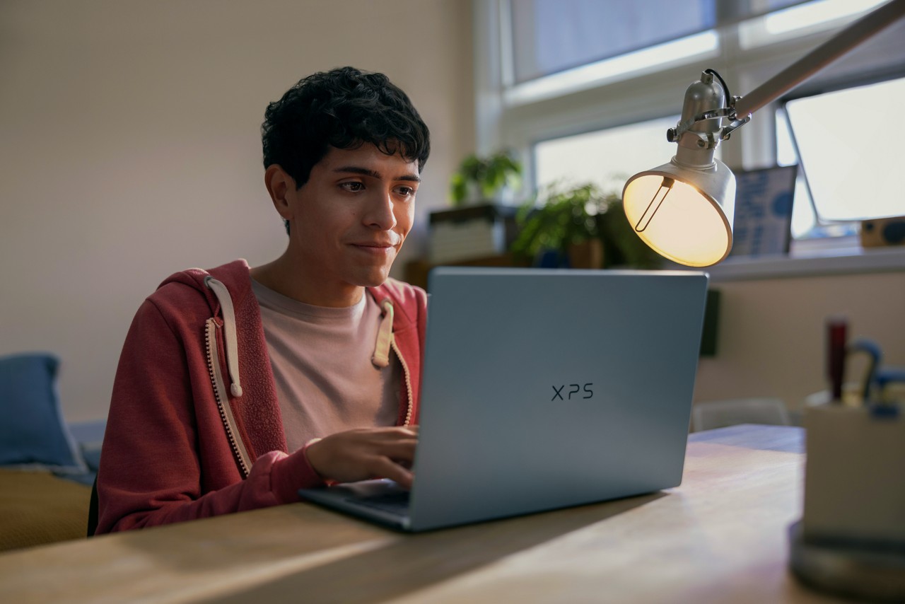 Student using a Dell XPS laptop at a desk under a lamp in a cozy study environment.