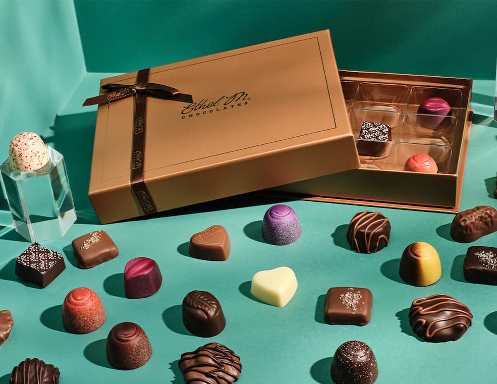 Ethel M Chocolates Copper Custom Box with a variety of chocolate gems surrounding it on a green background.