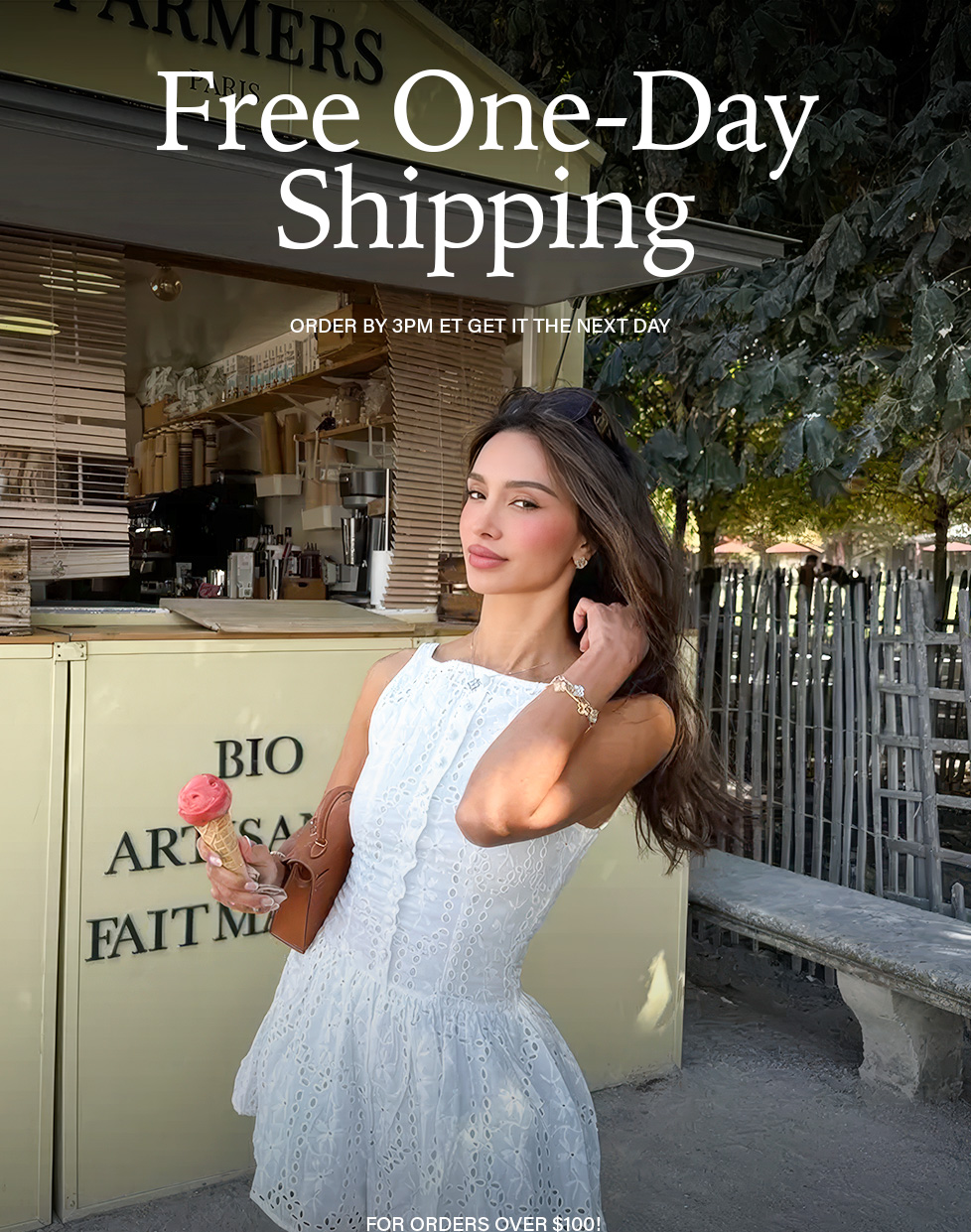 Smiling woman in white eyelet summer dress holds pink ice cream cone at an outdoor market, promoting Free One-Day Shipping over $100.