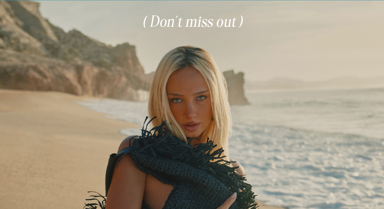 Blonde model wearing a dark, fringed beach top on a sandy shore with ocean and cliffs. Discover trendy coastal fashion – don't miss out!