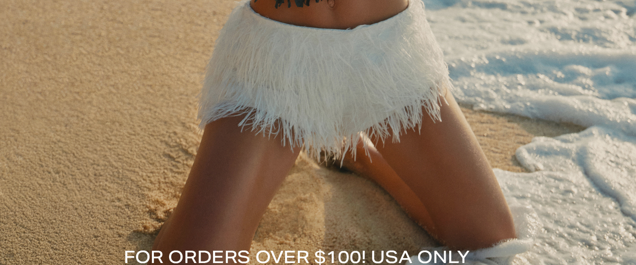 Person kneeling on a sandy beach wearing a white fringe swim bottom as ocean waves wash ashore. Shop stylish resort wear and get free shipping!