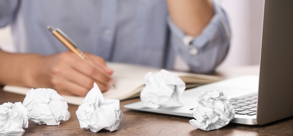 Image of a person writing with a gold pen in a notebook with scrunched up balls of paper littered across their desk
