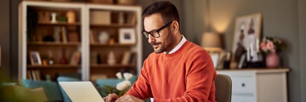 Image of a writer wearing an orange jumper and black glasses. He sits at his desk typing on his laptop.