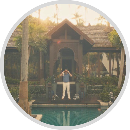 Image of a fancy tropical hotel surrounded by palm trees. An wealthy man adjusts his sunglasses overlooking a tiled pond.