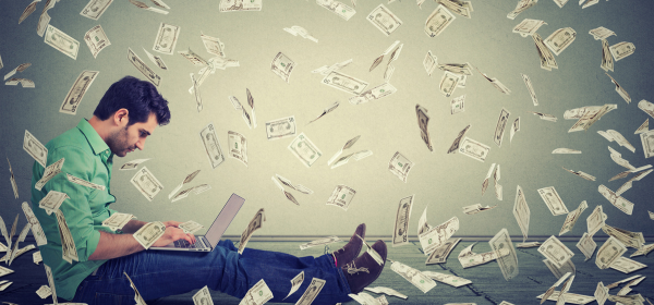 Image of a man writing on his laptop, seated on the floor with money raining downlls of paper littered across their desk
