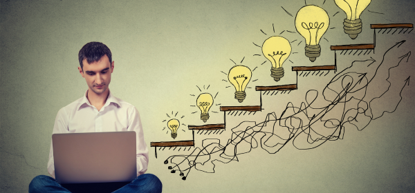 Man writing on his laptop. He sits on the floor leaning against a wall. On the wall, a staircase has been drawn with light bulbs placed on each step, increasing in size as it increases in height-1