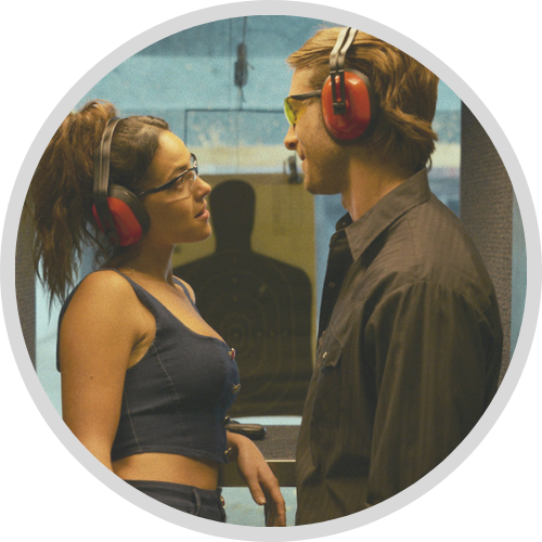 Glen Powell and Adria Arjona look at each other wearing red ear protectors at an indoor gun range in Hit Man