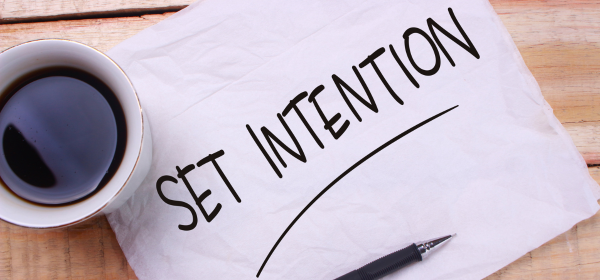 Image of a piece of paper that says Set Intention across it. The page lays on a wooden table next to a cup of coffee