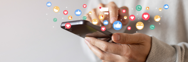 A hand positioned holding a phone with various social media emojis rising from it