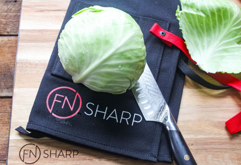 F.N. Sharp Santoku Knife and apron on Acacia wood cutting board with a head of cabbage
