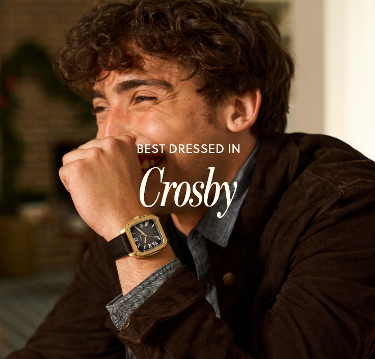 Young man laughing, wearing a Fossil Crosby watch with black leather strap, gold case, and black dial” set against a dark festive background.