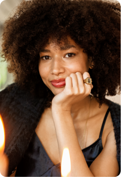 Close-up portrait of a woman with curly dark hair, faint smile, and hand on chin, wearing a black top and a Fossil ring watch on her left hand