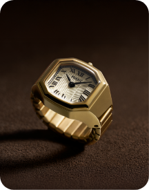 A close-up of a gold-toned watch ring with an octagonal case and a white textured dial, set against a dark background