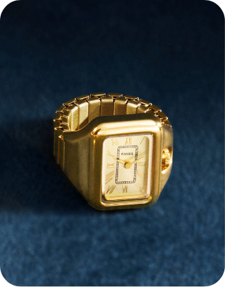 A close-up of a gold-toned watch ring with a rectangle case and a cream dial, set against a dark background