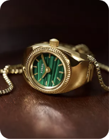 A close-up of a gold-toned watch ring with a circular case and a green dial attached to a gold chain