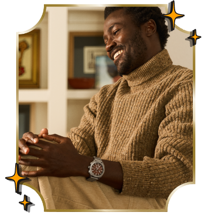 Man sitting and smiling while looking off-camera, hands on his knee, wearing a cozy brown turtleneck and a Fossil red dial watch