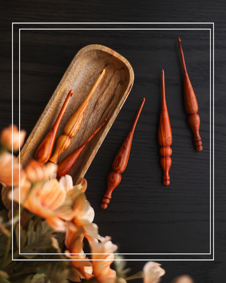 wood alpha crochet hooks and a wood hook tray are arranged on a black table underneath peach flowers