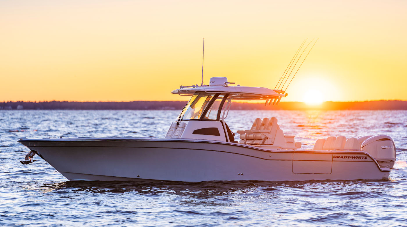 A sleek, blue center console fishing boat floating on calm water at sunset. The bright sun is low on the horizon, casting a warm golden glow across the sky and reflecting on the gentle waves. The boat features a T-top with fishing rods angled upward, three seats at the helm, and outboard motors at the stern.