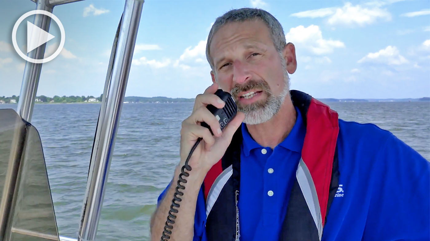 Man at the helm of a boat holding a VHF radio microphone to his mouth.