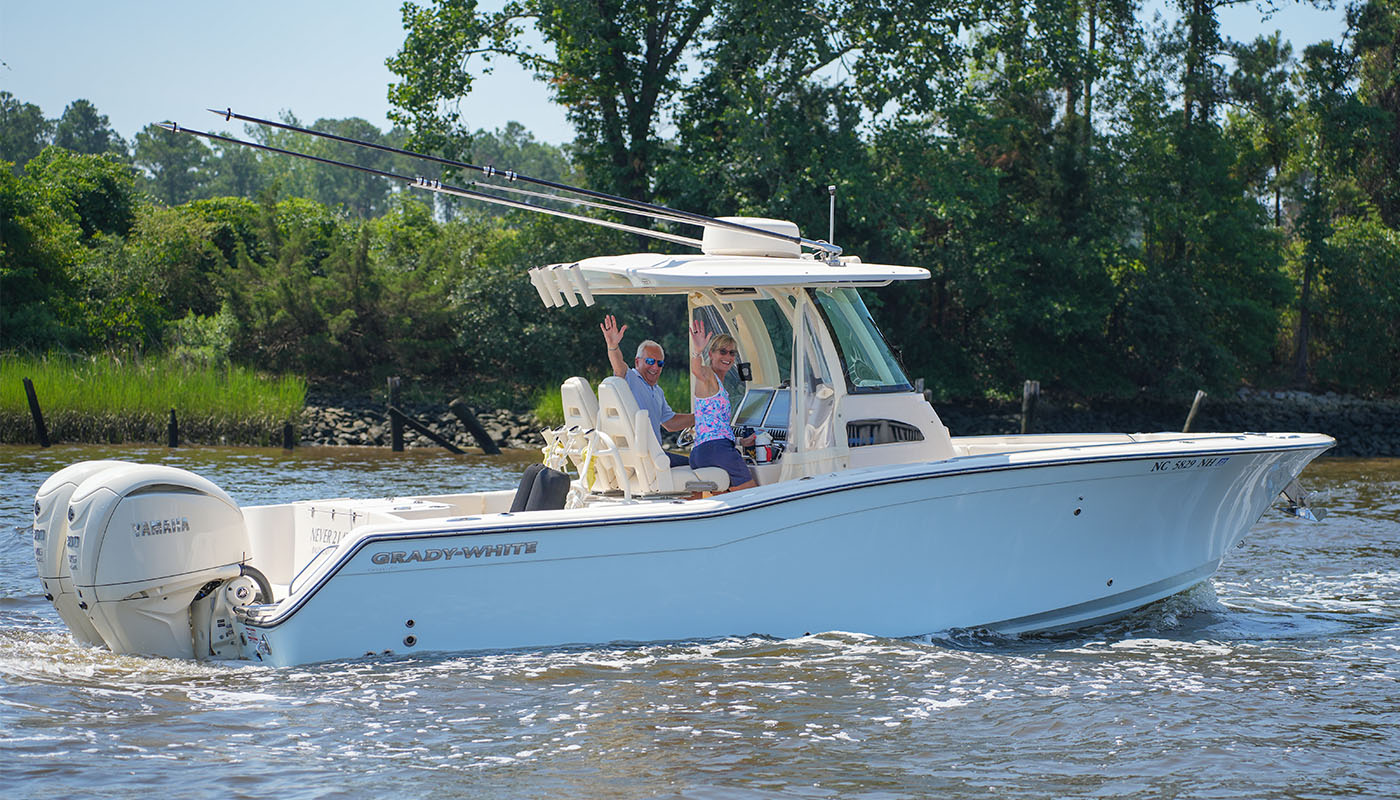 Two people smiling and waving from the helm of a Grady-White Canyon 271 center console boat as it cruises on the water near a tree-lined shoreline.