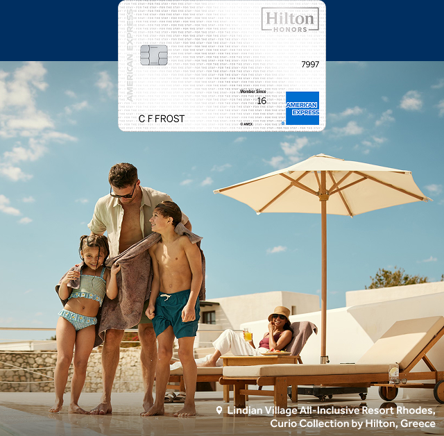 A Hilton Honors Amex card sits above a family in swimwear at Lindian Village All-Inclusive Resort Rhodes, Curio Collection by Hilton, Greece.
