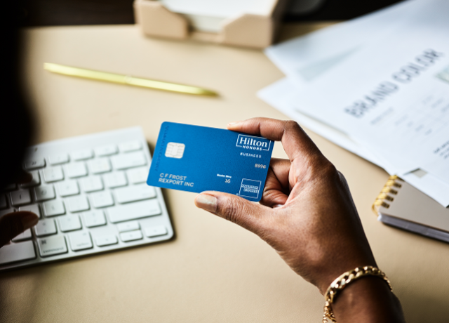 Person holding a blue Hilton Honors Amex business card at a desk.