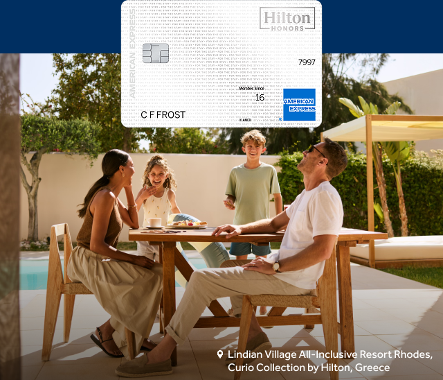 A Hilton Honors Amex card sits above a family laughing at a table by a pool at Lindian Village All-Inclusive Resort Rhodes, Curio Collection by Hilton, Greece.