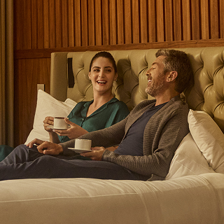 Two people sit on a bed with a tufted headboard, holding cups, with wooden paneling in the background.