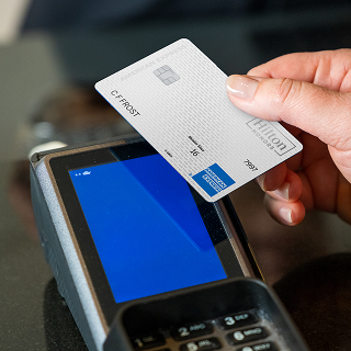 A hand holds a Hilton Honors Amex card near a payment terminal for contactless payment.