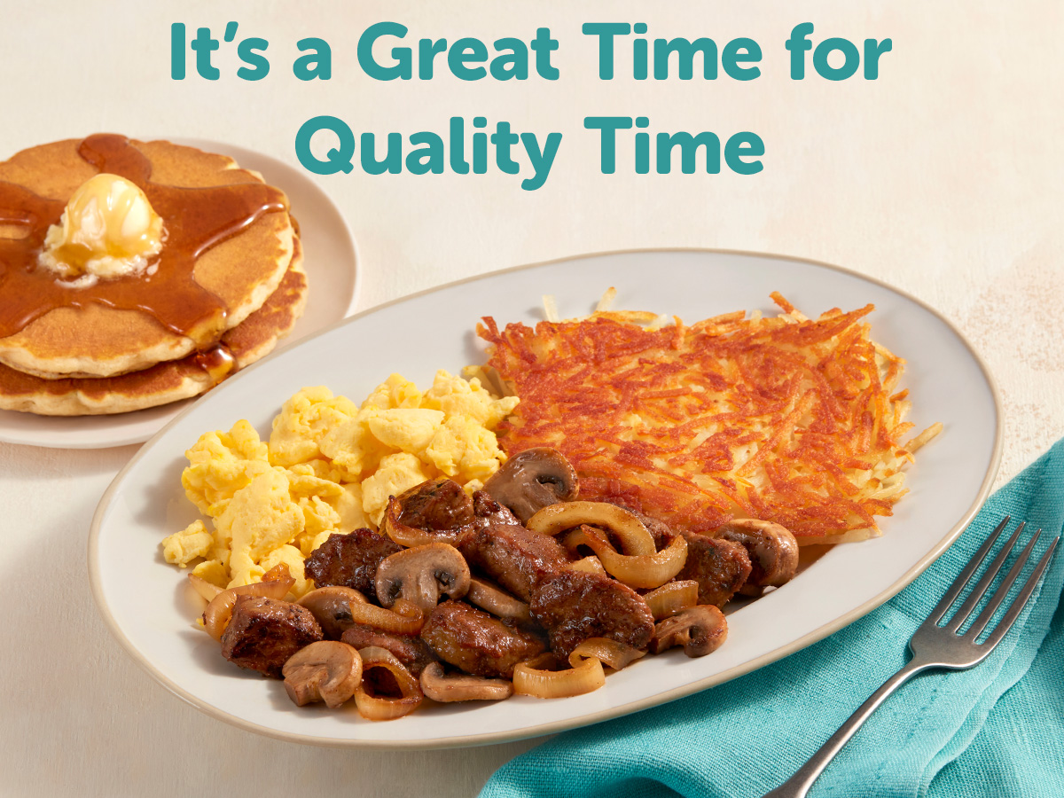 Two pancakes topped with syrup and butter, with a plate of sides including mushrooms, scrambled eggs and hash browns.