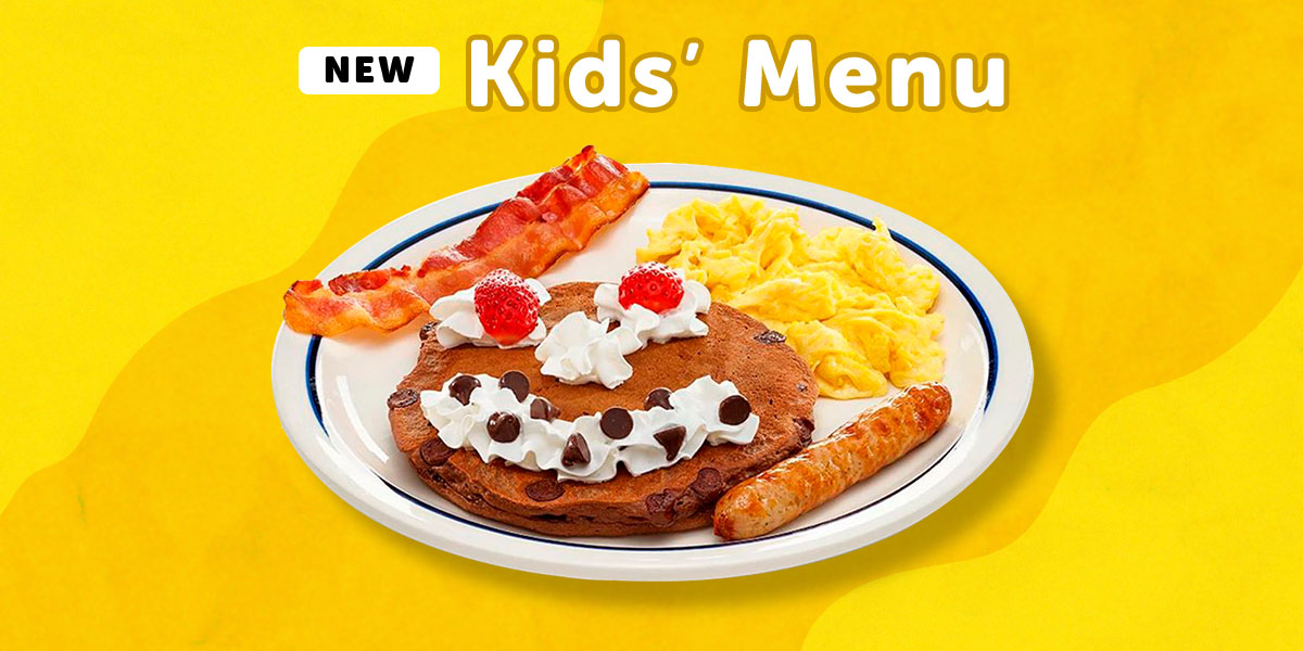 The Happy Face Combo from IHOP’s Kids’ Menu, featuring scrambled eggs, a bacon strip, a sausage link, and a pancake with a face: Maraschino cherry eyes and a whipped topping nose and mouth with some chocolate chips for teeth.
