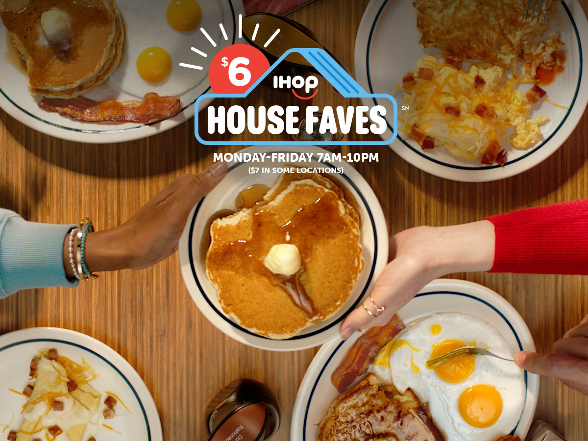 Two different women’s hands hold a plate of buttermilk pancakes over a table from opposite sides. On the table are IHOP’s House Faves meals, including the Breakfast Faves Combo and Ham and Cheese Omelette.