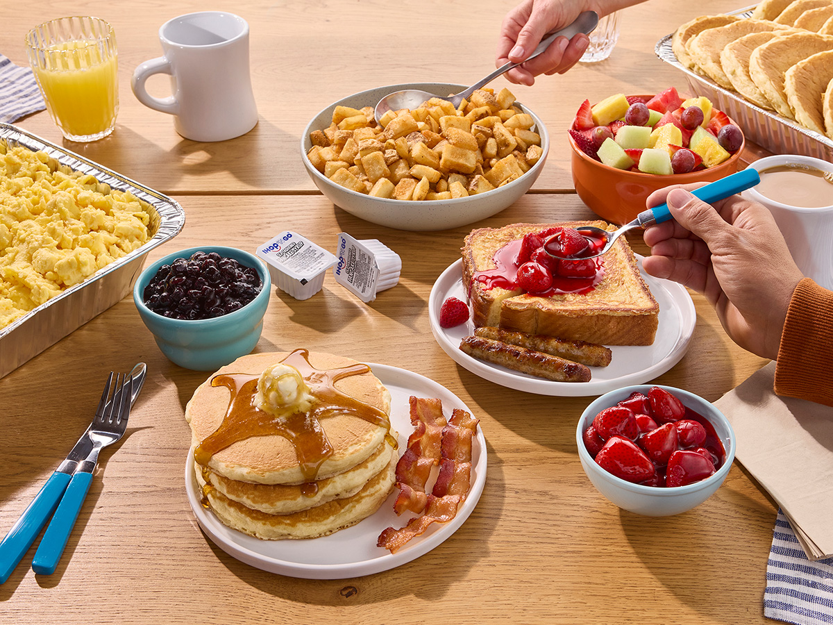 A full spread of IHOP Catering on a table, including tin trays filled with scrambled eggs and pancakes, a plate of pancakes and bacon, cups of fruit, and French Toast
