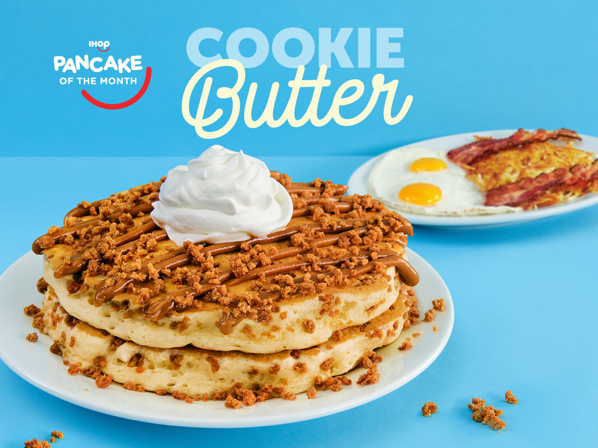 A shot of two Cookie Butter Pancakes, topped with Biscoff® Cookie Butter, cookie pieces, and whipped cream, with a plate of eggs sunny side up, hash browns, and bacon strips in the background.