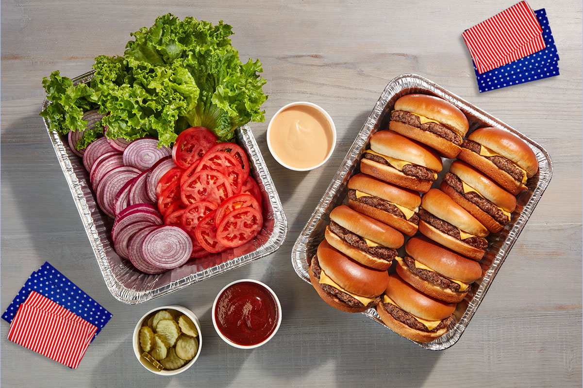 Two catering tins. One filled with IHOP Steakburgers and the other filled with fixins’ like lettuce, tomato, and onions. A cup of pickles, ketchup and spread are also on the table, along with American flag inspired napkins.