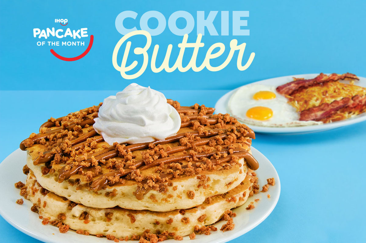 An image of Cookie Butter Pancakes, topped with cookie crumbles and cookie butter, with a plate of eggs, bacon, and hashbrowns in the background.