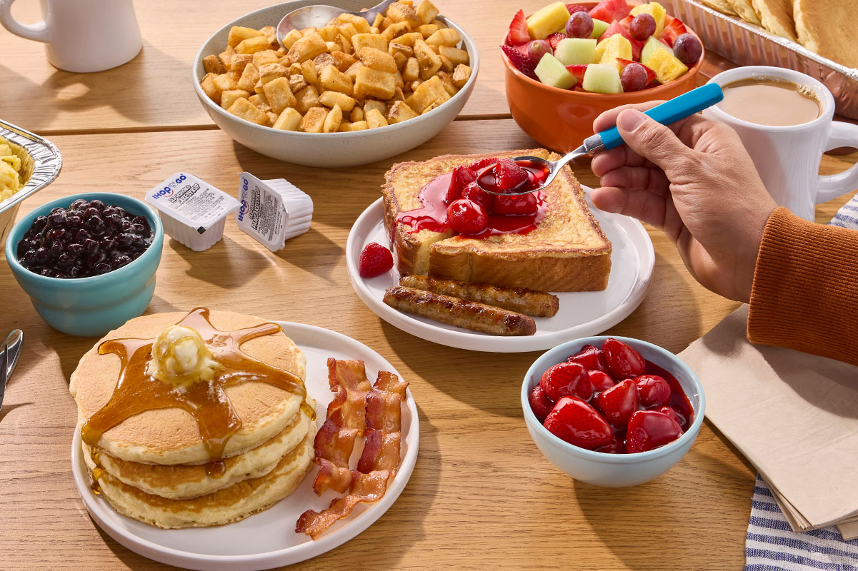 A table with IHOP Catering on it, including trays of pancakes and scrambled eggs. Hands reach in with forks to plates of French Toast and potatoes.