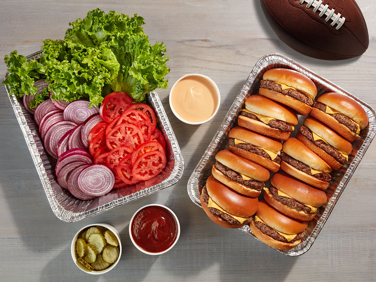 Two catering trays, one with IHOP Steakburgers and one with fixings, are on a table, along with a football and cups of condiments.