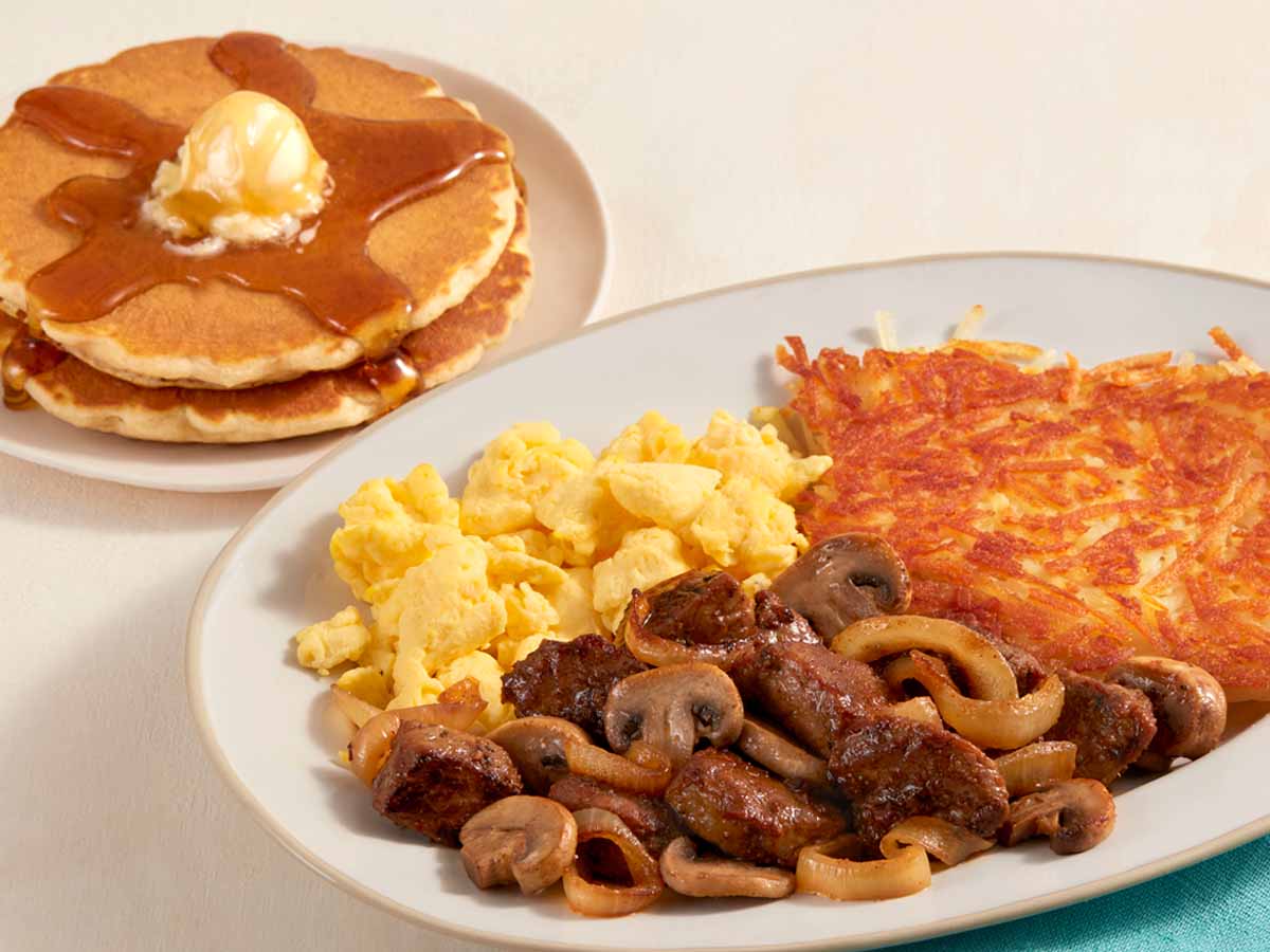 A plate of scrambled eggs, hash browns, steak tips and mushrooms, with a plate of two buttermilk pancakes with syrup and butter.