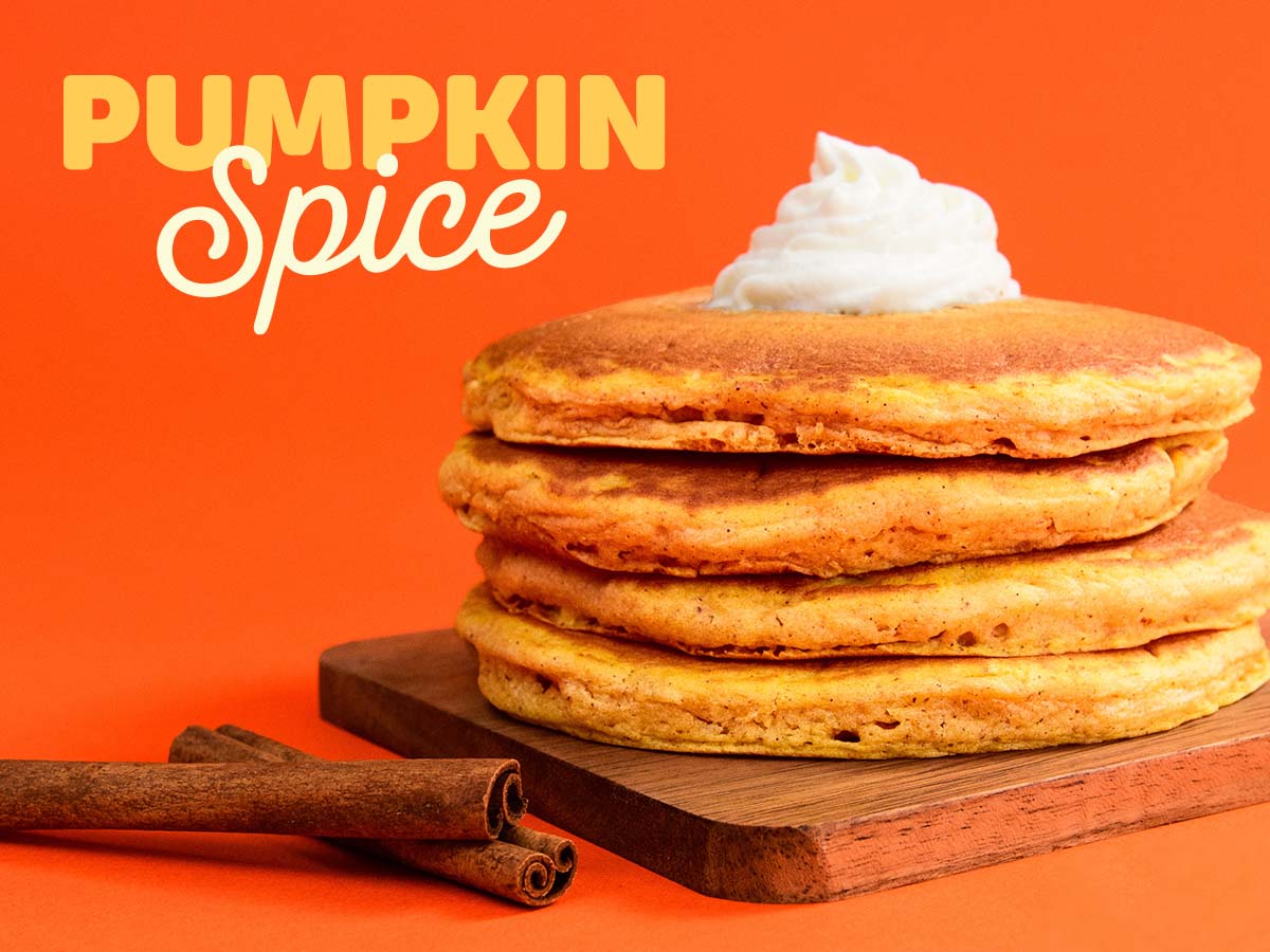 A Full Stack of Pumpkin Spice Pancakes with whipped topping on a wood cutting board, with two sticks of cinnamon beside it, against an orange background.
