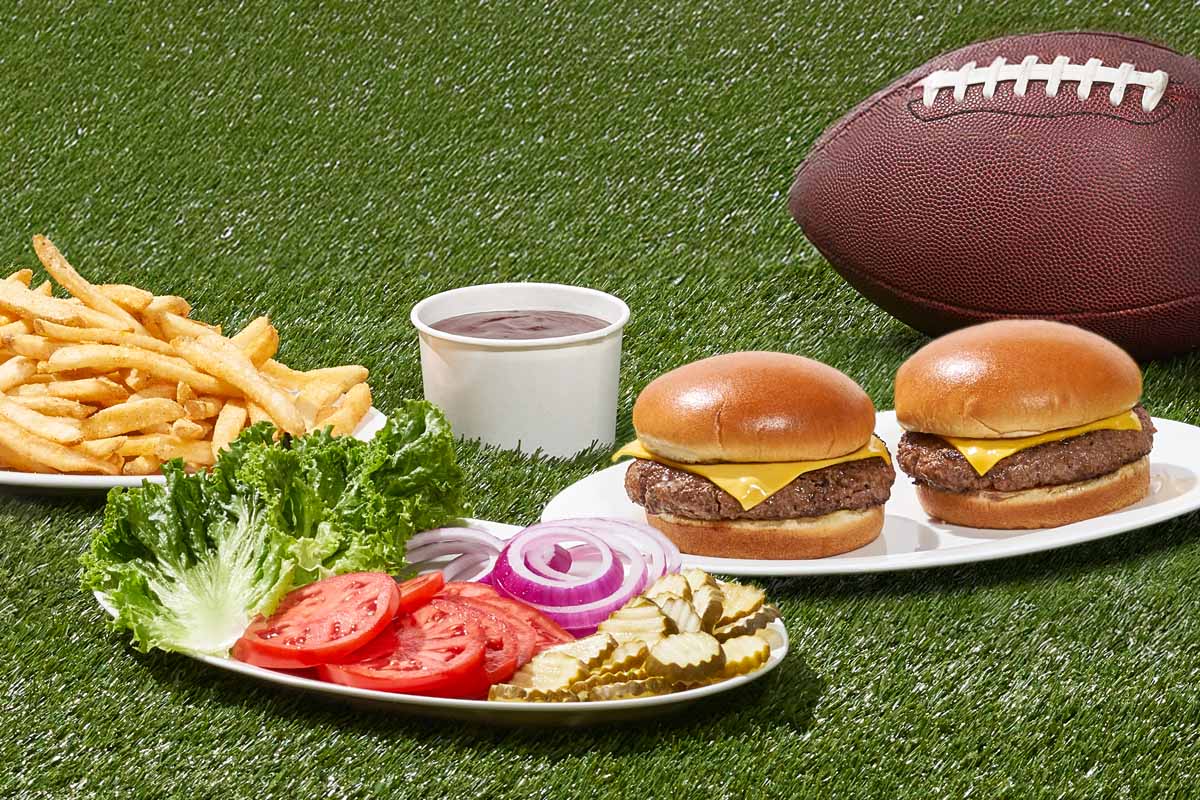 Burgers with fixings and fries on plates, set on top of green turf. A football is in the background