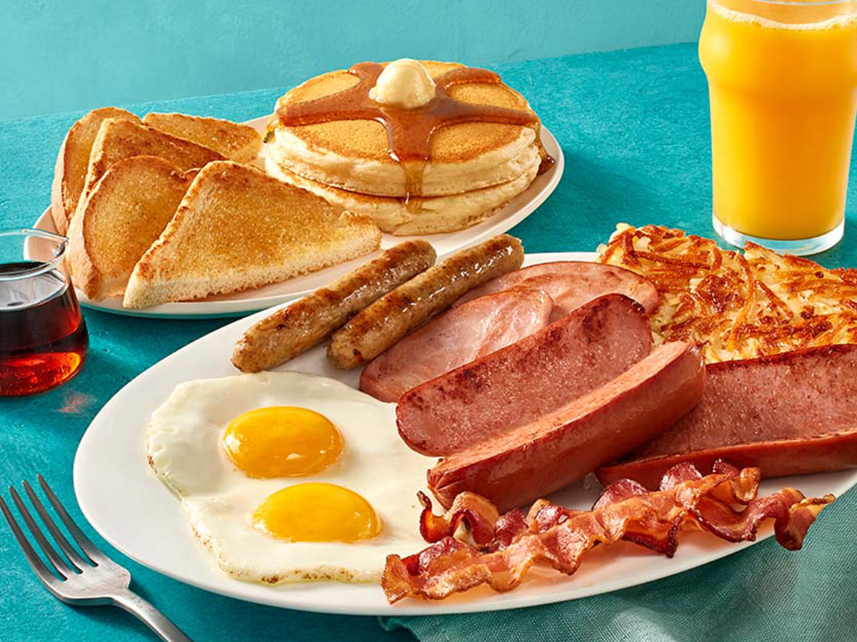 A plate with two eggs, hash browns, two slices of ham, two bacon strips, two pork sausage links, and two smoked sausage links. A second plate has two buttermilk pancakes and toast.